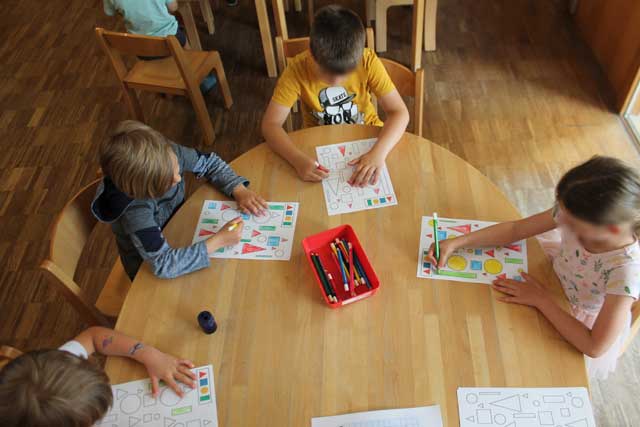 Kinder sitzen an einem Tisch und malen mit Buntstiften verschiedenen Formen aus