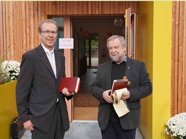 Pfarrer Eberhardt und Kirchensteiner stehen vor den Eingangstür zur neuen Holzmodulanlage